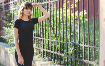 A woman in a black dress leans against a wrought iron fence, surrounded by green foliage. She has her right hand resting on the fence, looking thoughtfully into the distance. The setting appears to be outdoors on a sunny day.