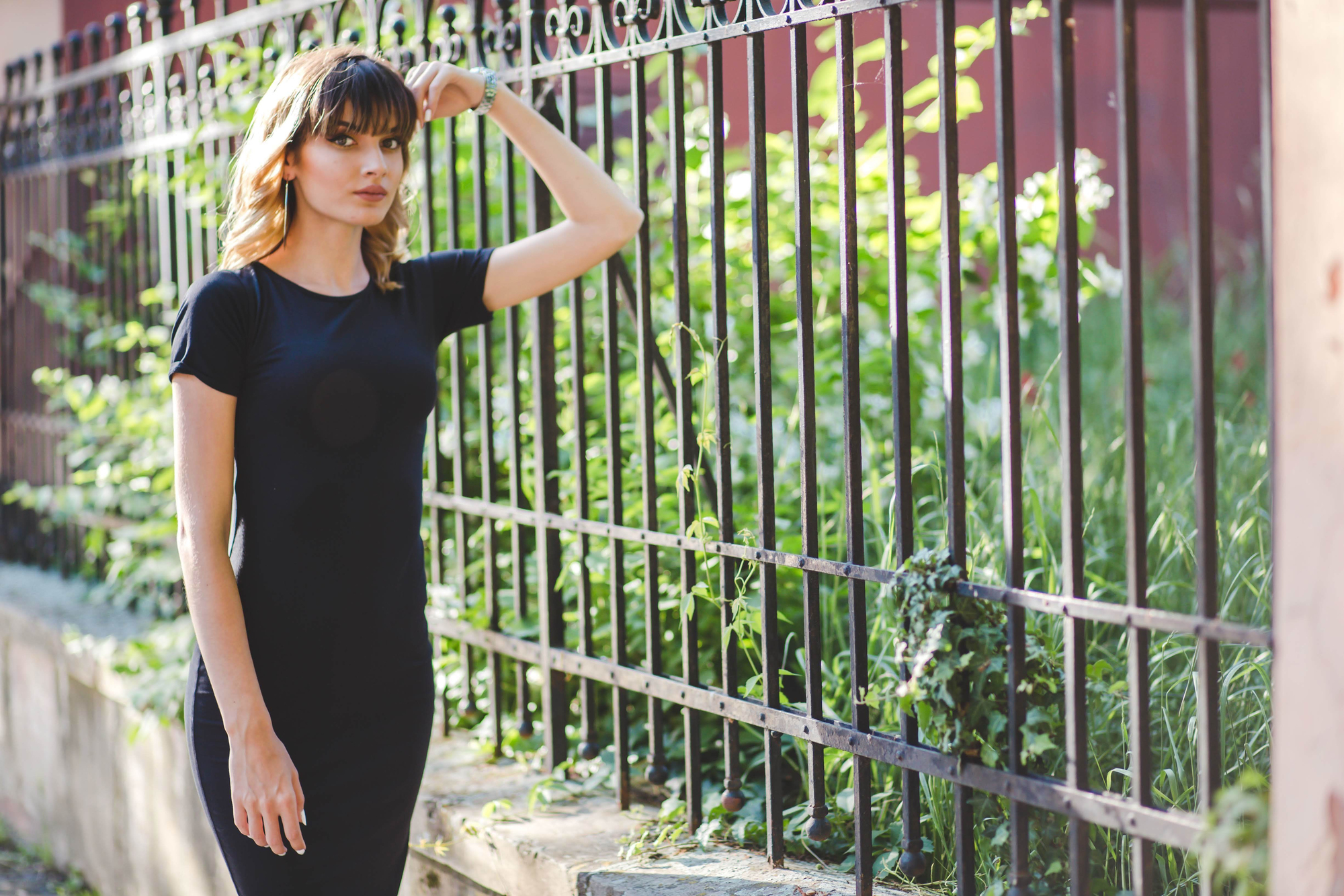 A woman in a black dress leans against a wrought iron fence, surrounded by green foliage. She has her right hand resting on the fence, looking thoughtfully into the distance. The setting appears to be outdoors on a sunny day.