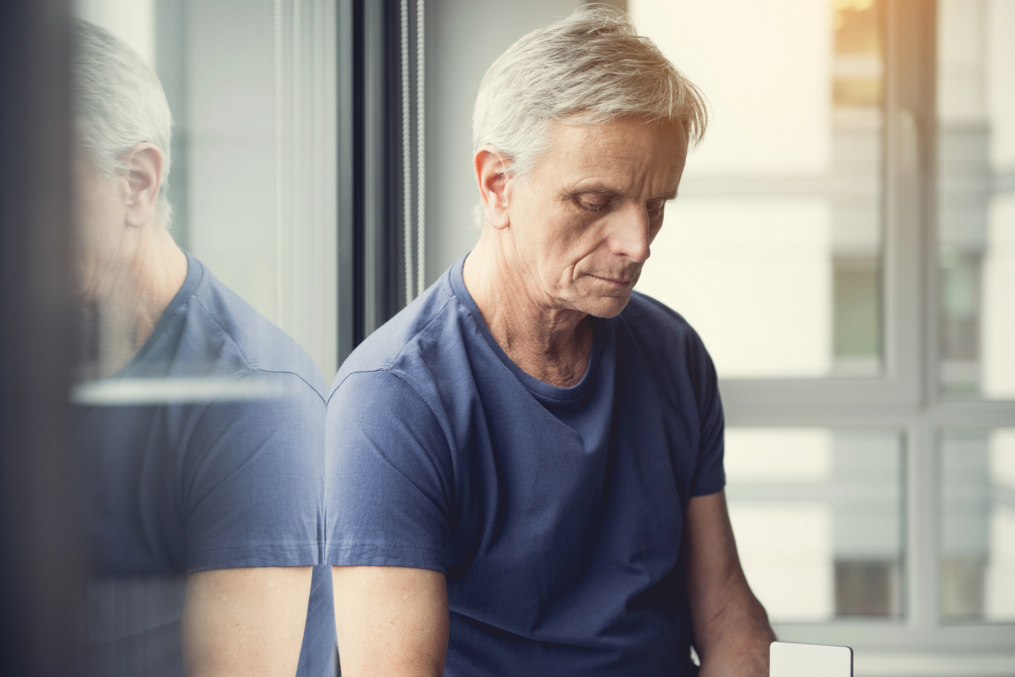 Older man sitting near a window, wearing a blue shirt, and looking downward. A reflection of the man is visible in the glass. The scene is softly lit with natural light from outside.