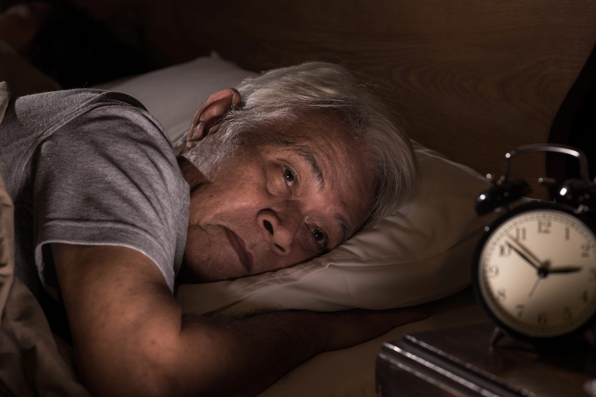 An elderly man with gray hair is lying in bed, propped on a pillow, and looking past the camera with a thoughtful expression. An alarm clock showing 4:15 is on a nearby table, dimly lit by a soft light.