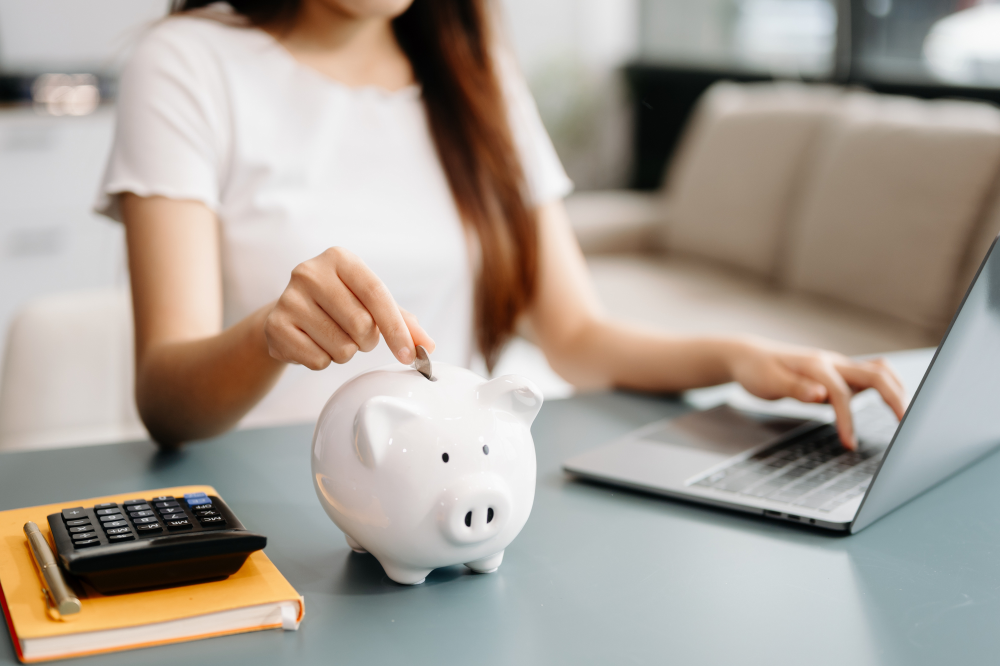 A person places a coin into a white piggy bank while working on a laptop. A calculator and a notebook are visible on the table. The setting appears to be a modern, bright living room or office.