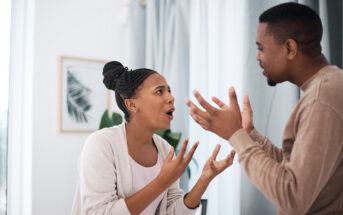 A woman and a man are standing indoors, engaged in a heated discussion. They are facing each other with expressive gestures. The room is softly lit, with a framed picture and a plant in the background.