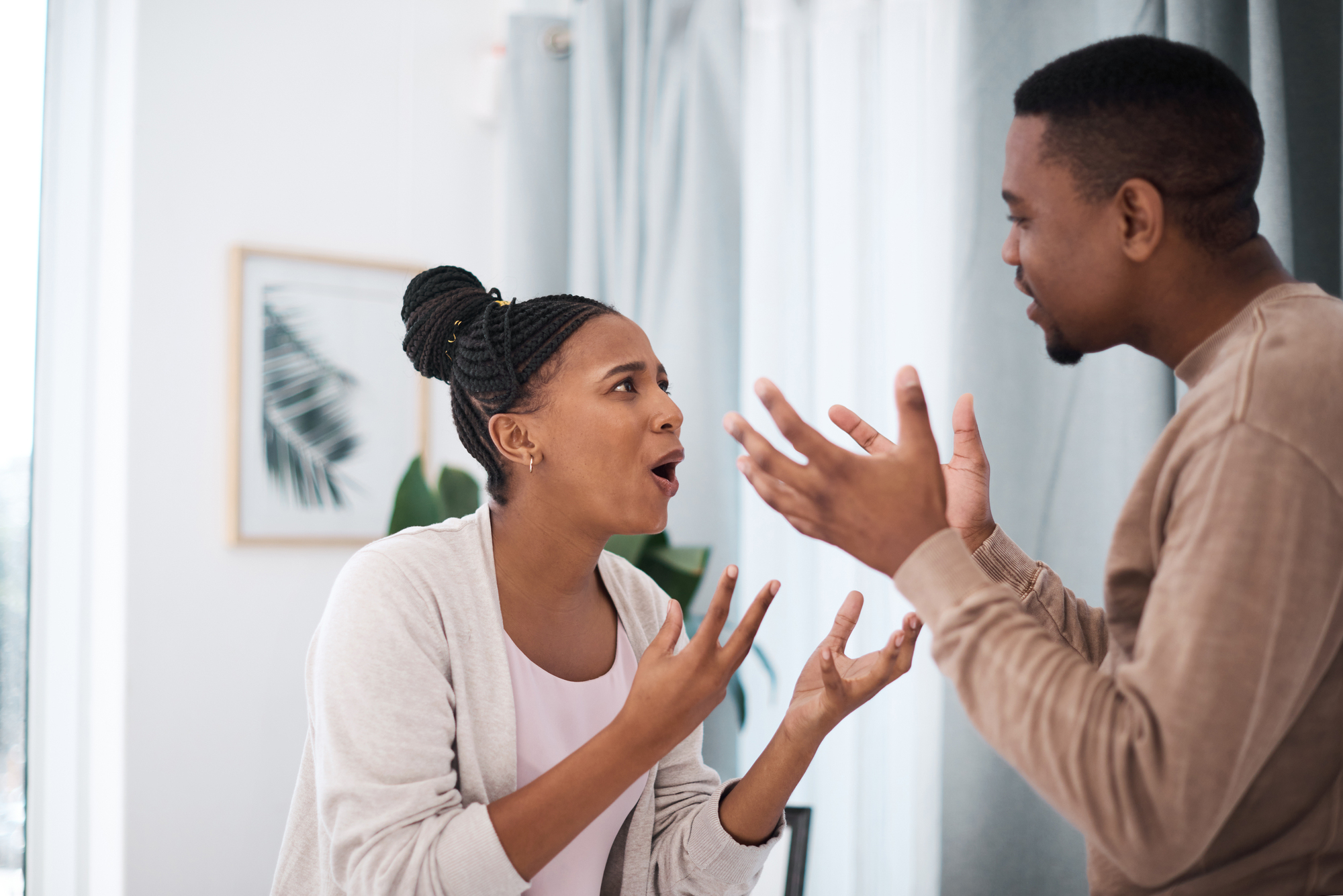 A woman and a man are standing indoors, engaged in a heated discussion. They are facing each other with expressive gestures. The room is softly lit, with a framed picture and a plant in the background.