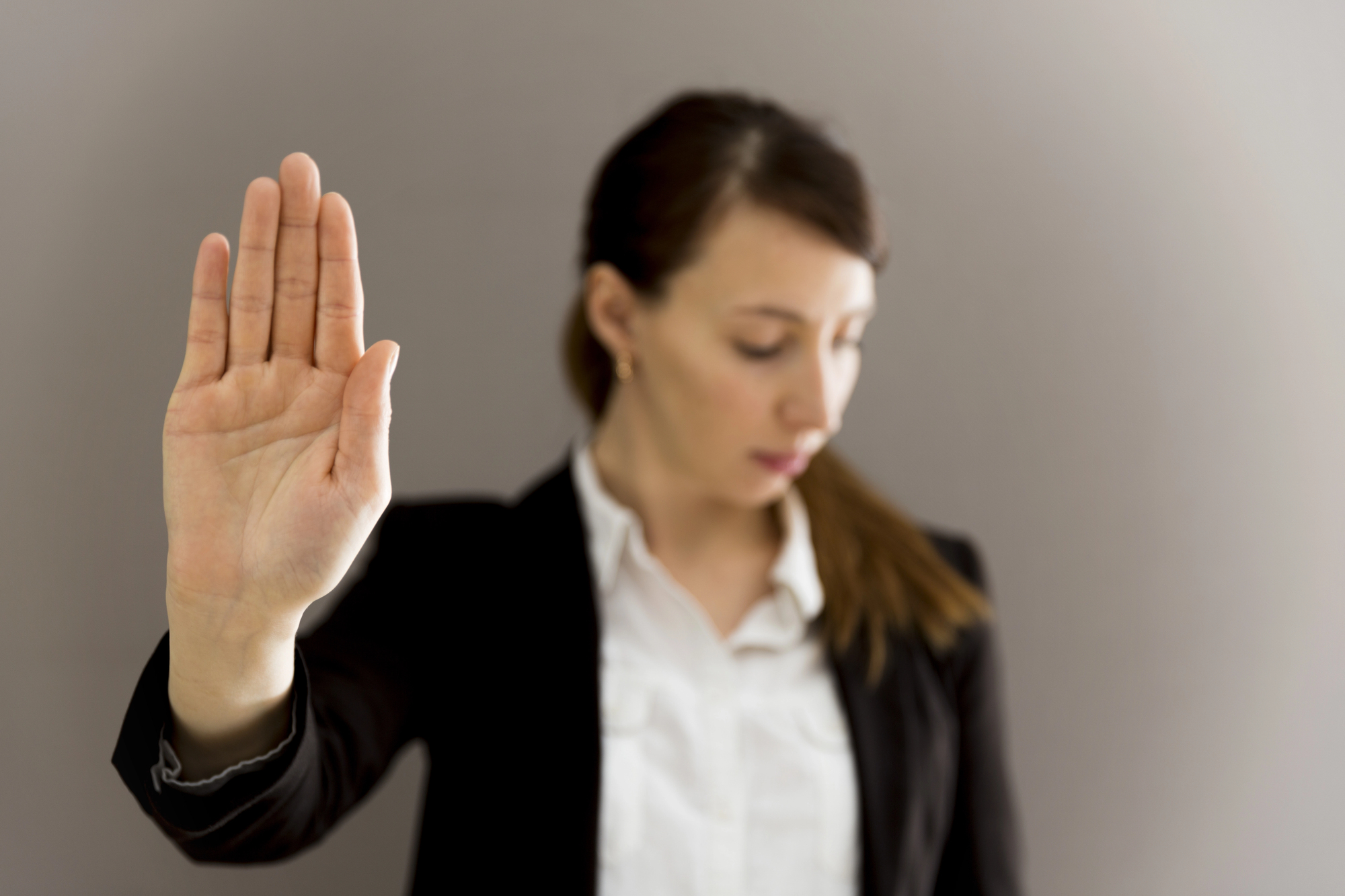 A woman in a black blazer and white shirt holds up her hand with palm facing outward, as if signaling to stop. She is looking downward, standing against a plain gray background.