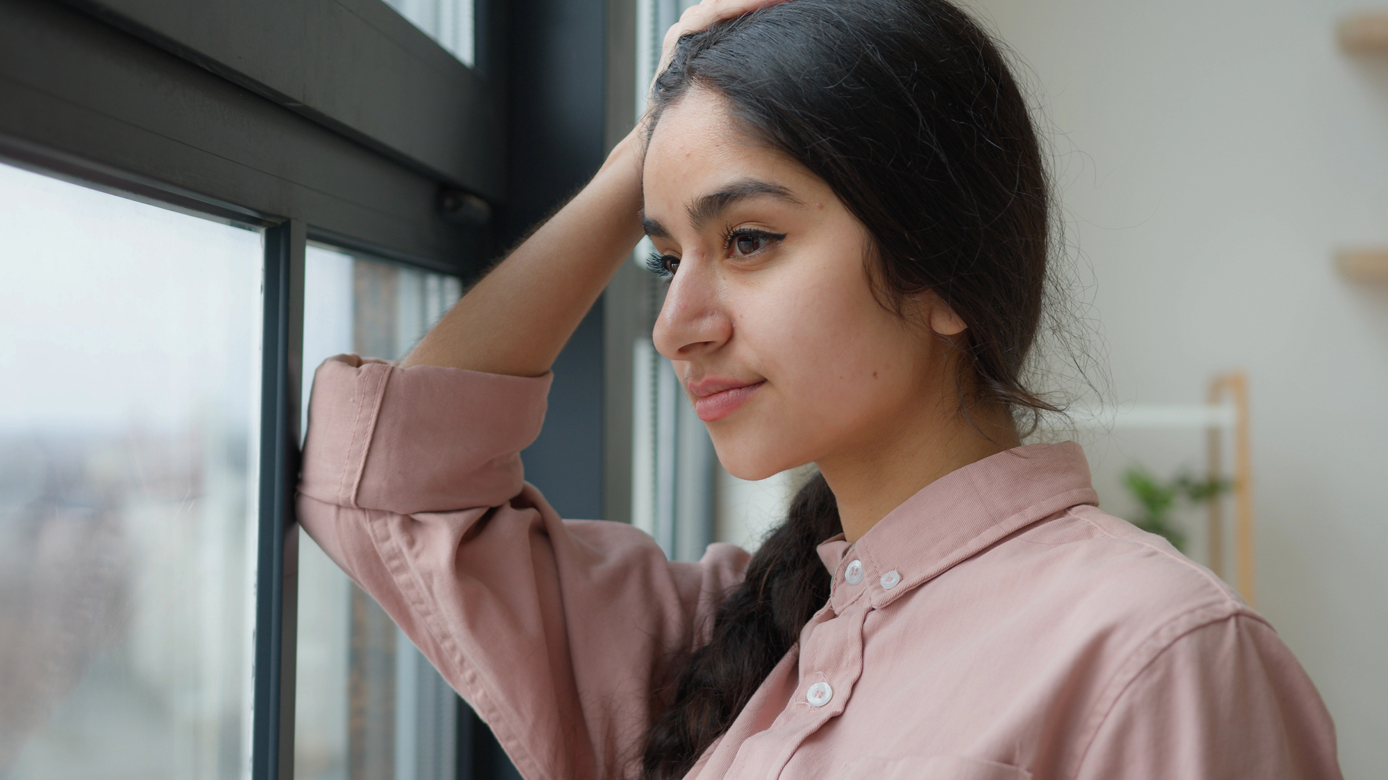 A person with long dark hair in a braid leans against a window, looking outside with a thoughtful expression. They wear a pink button-up shirt and rest one hand on their head. The background is softly blurred.