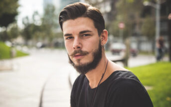 A man with a beard and mustache sits outdoors, looking at the camera. He is wearing a black T-shirt and a necklace. The background is blurred, showing trees and buildings in an urban setting.