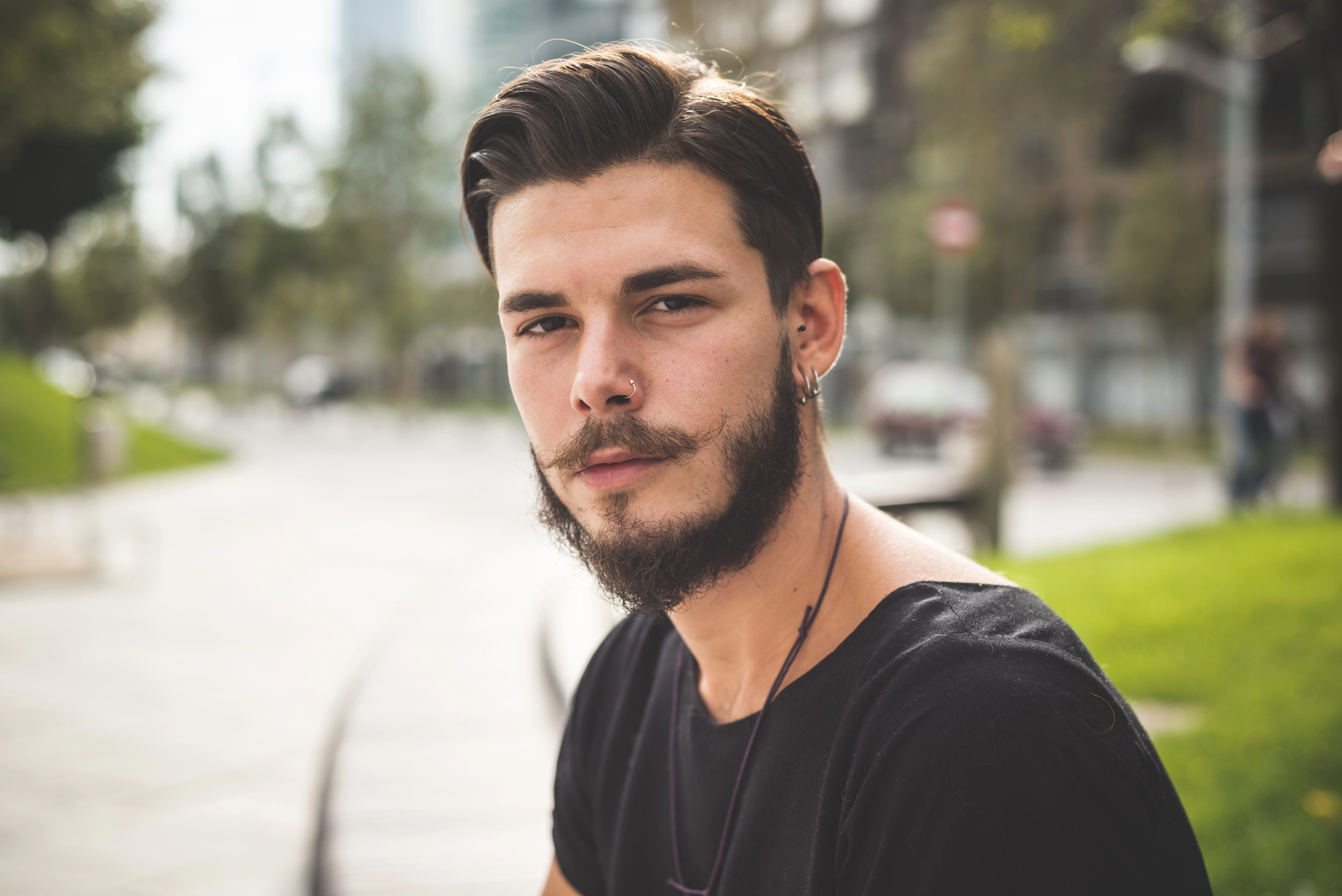 A man with a beard and mustache sits outdoors, looking at the camera. He is wearing a black T-shirt and a necklace. The background is blurred, showing trees and buildings in an urban setting.