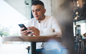 A man in a white shirt is sitting at a cafe table, focused on his smartphone. A cup of coffee is on the table. The cafe interior is visible, and daylight streams through the window, highlighting street activity outside.