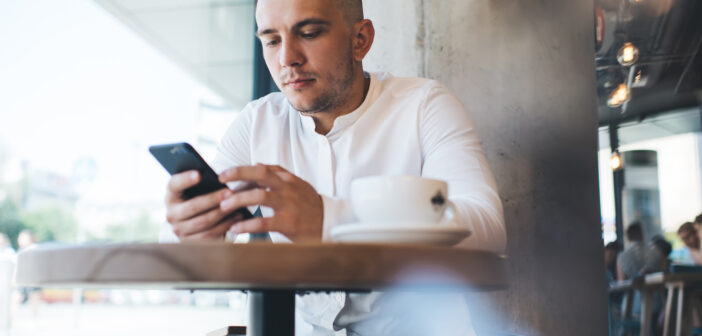 A man in a white shirt is sitting at a cafe table, focused on his smartphone. A cup of coffee is on the table. The cafe interior is visible, and daylight streams through the window, highlighting street activity outside.