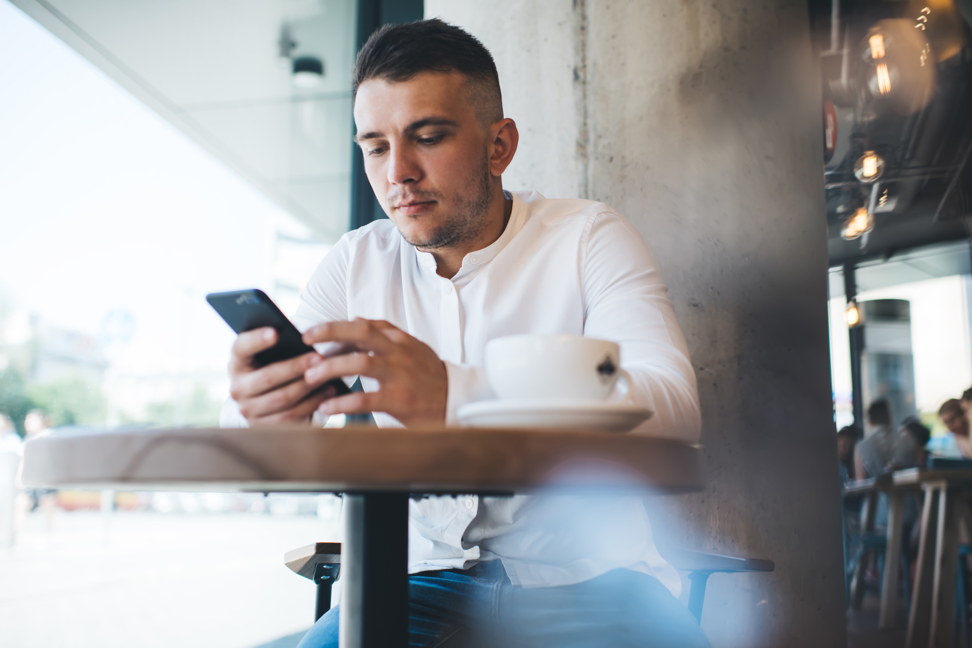 A man in a white shirt is sitting at a cafe table, focused on his smartphone. A cup of coffee is on the table. The cafe interior is visible, and daylight streams through the window, highlighting street activity outside.