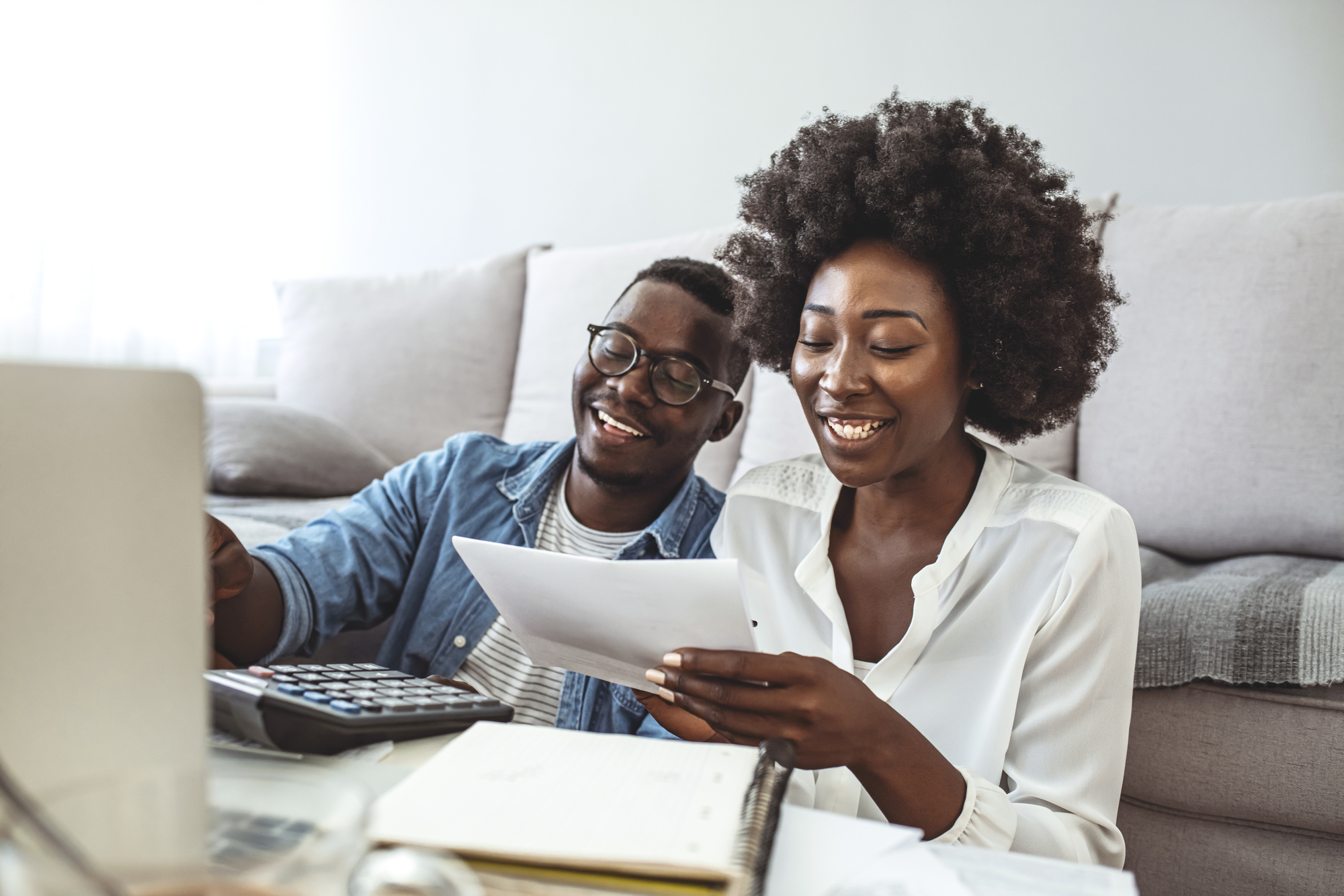 A smiling couple sits together on the floor in a living room, engaged in reviewing documents. The man holds a calculator, and the woman holds a paper. A laptop and notebook are visible on the table next to them.