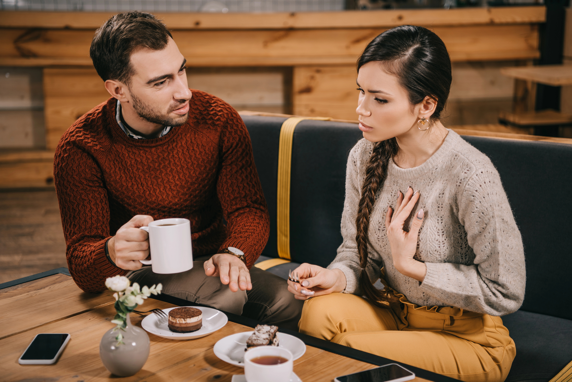 A man and woman sitting in a cozy cafe, engaged in conversation. The man holds a mug, and there's a dessert on the table. The woman gestures with her hand. They seem to be having a serious discussion. Smartphones and a vase with flowers are on the table.