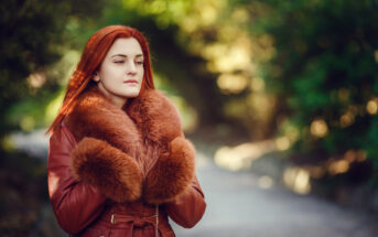 A woman with red hair stands outdoors, wearing a warm coat with a large fur collar. The background is a blurred natural setting with green trees, and she appears thoughtful and serene.