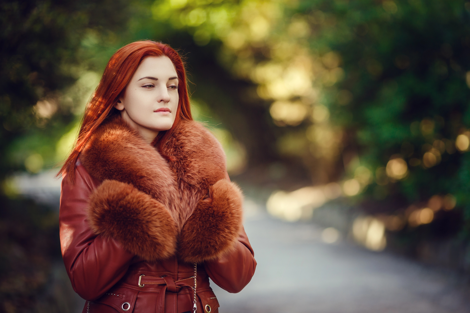 A woman with red hair stands outdoors, wearing a warm coat with a large fur collar. The background is a blurred natural setting with green trees, and she appears thoughtful and serene.