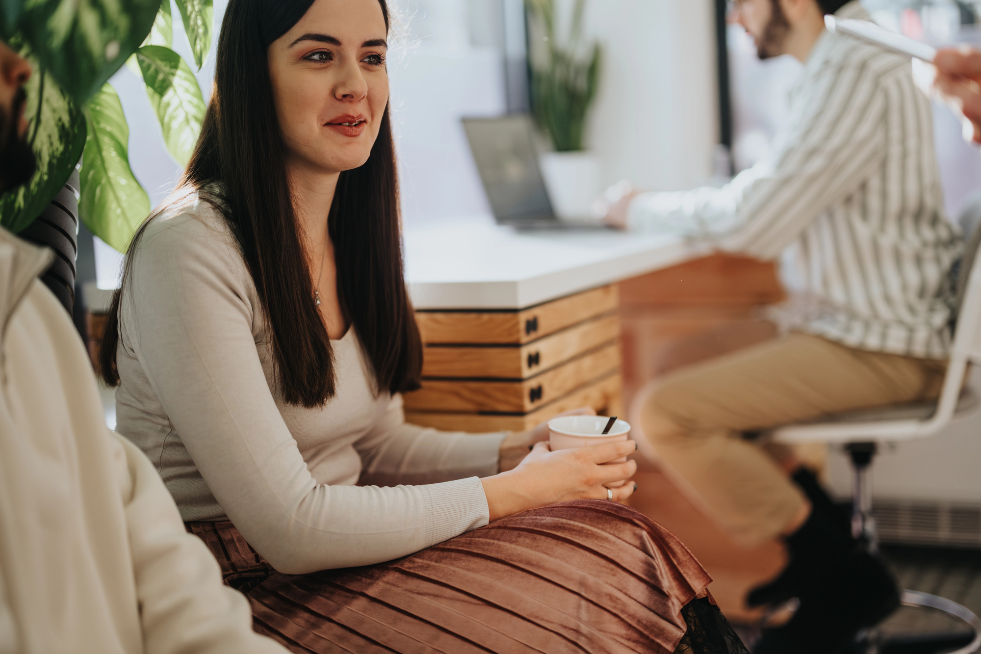 A woman with long dark hair, wearing a beige sweater and pleated skirt, sits in an office holding a cup. She is engaged in conversation. In the background, a man works on a laptop at a desk with a plant nearby.