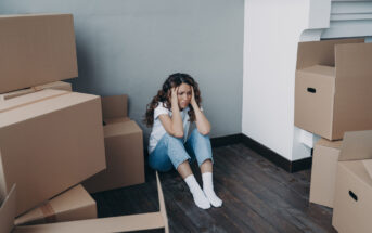 A woman sits on the wooden floor in a corner, looking distressed. She is surrounded by cardboard boxes. She wears a white shirt, blue jeans, and white socks, with her hands on her face. The walls are gray and white.