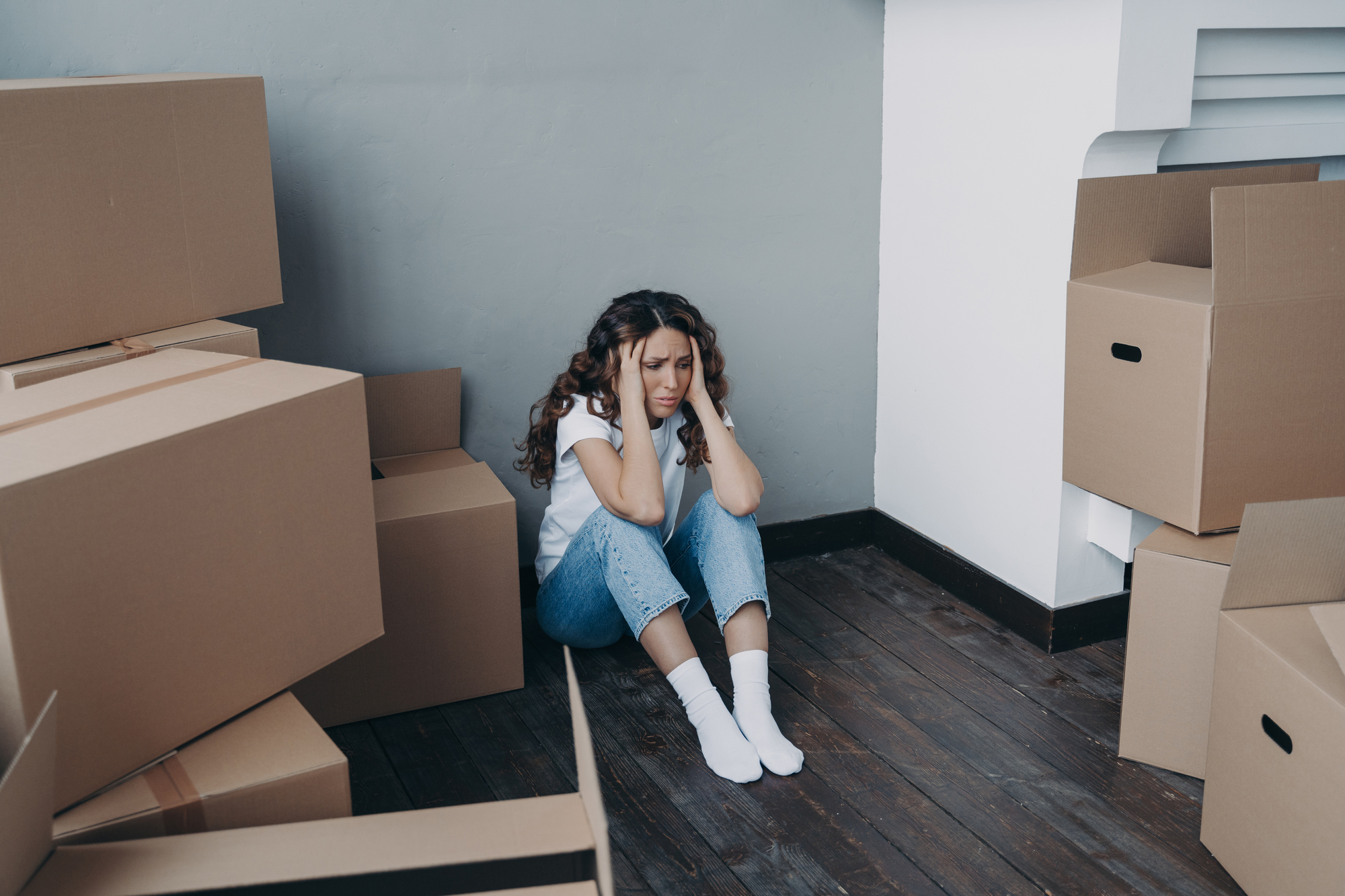 A woman sits on the wooden floor in a corner, looking distressed. She is surrounded by cardboard boxes. She wears a white shirt, blue jeans, and white socks, with her hands on her face. The walls are gray and white.