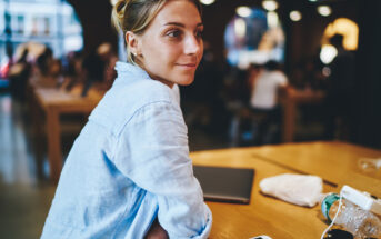 A woman with a light blue shirt sits at a wooden table in a bustling cafe. She has a slight smile and looks to the side. The table has a laptop, tablet, and a water bottle. People are blurred in the background, suggesting a lively atmosphere.