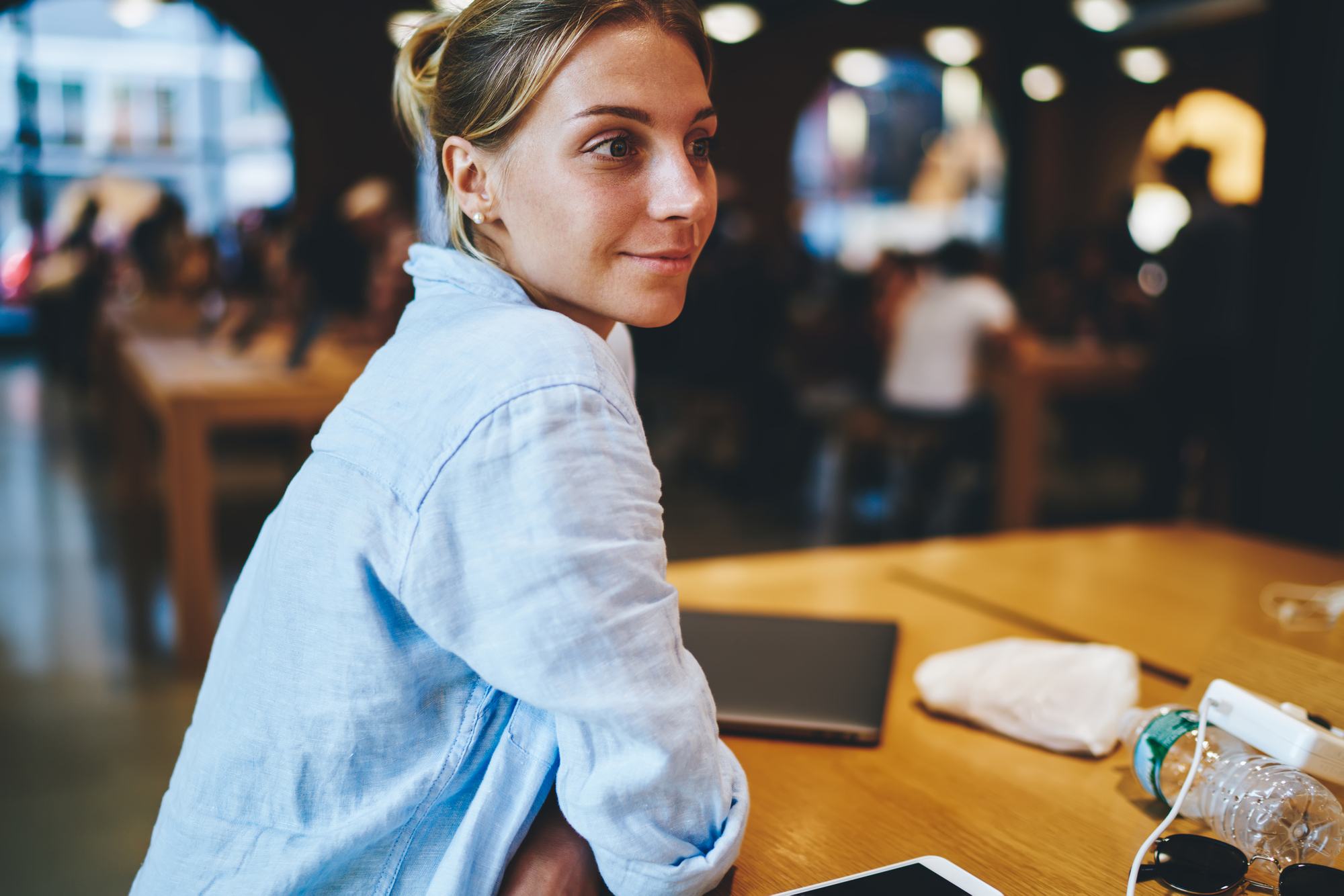 A woman with a light blue shirt sits at a wooden table in a bustling cafe. She has a slight smile and looks to the side. The table has a laptop, tablet, and a water bottle. People are blurred in the background, suggesting a lively atmosphere.