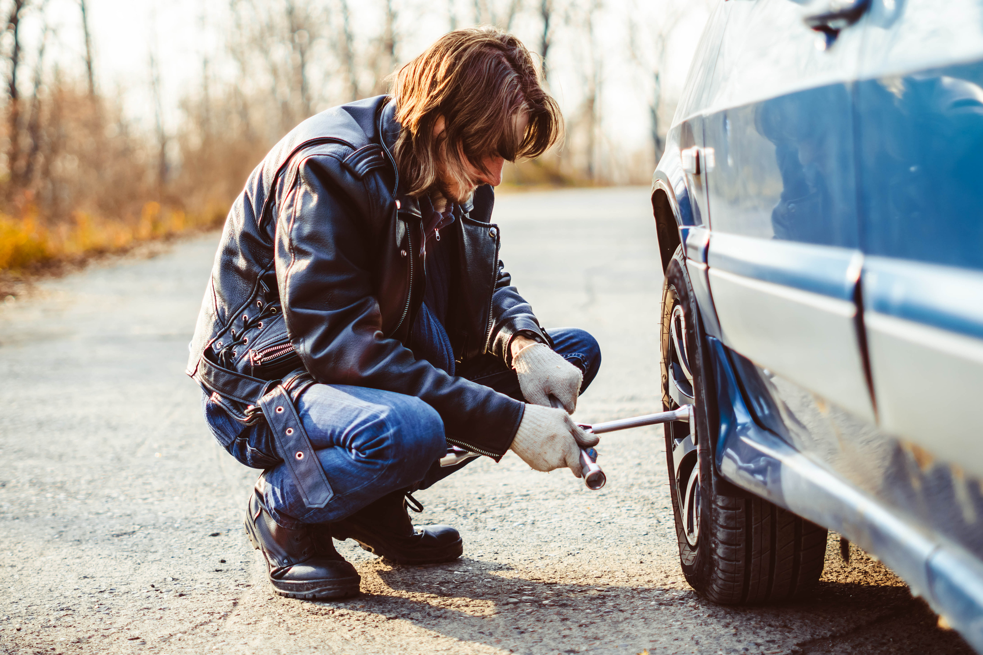 A person in a leather jacket and gloves is crouched down on a road, using a wrench to adjust the wheel of a parked car. The background shows a blurred view of trees and a clear sky.