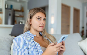 A woman with long blonde hair, wearing a blue and white striped shirt, is sitting on a sofa holding a smartphone. She gazes thoughtfully into the distance in a modern living room setting.