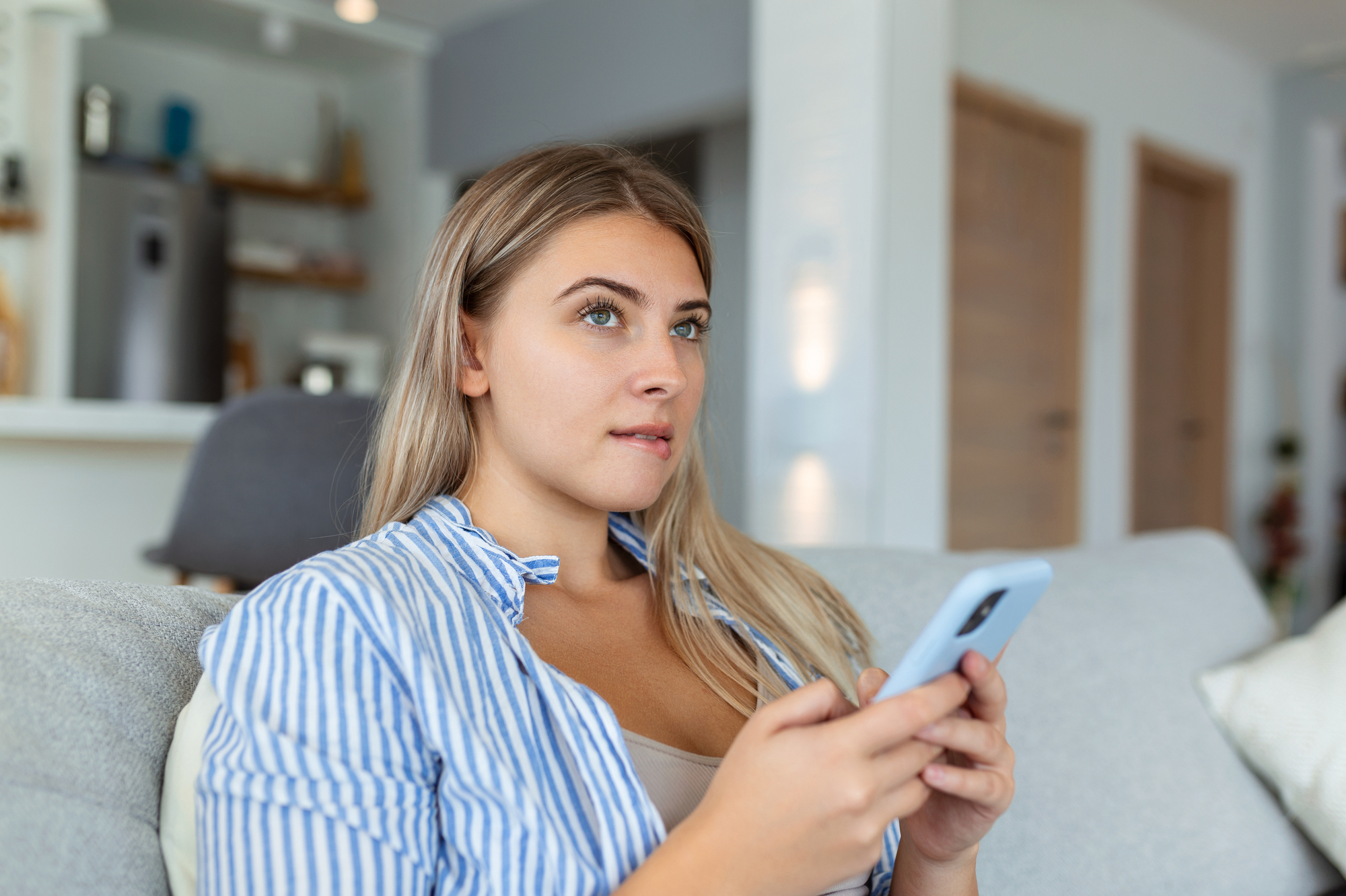 A woman with long blonde hair, wearing a blue and white striped shirt, is sitting on a sofa holding a smartphone. She gazes thoughtfully into the distance in a modern living room setting.