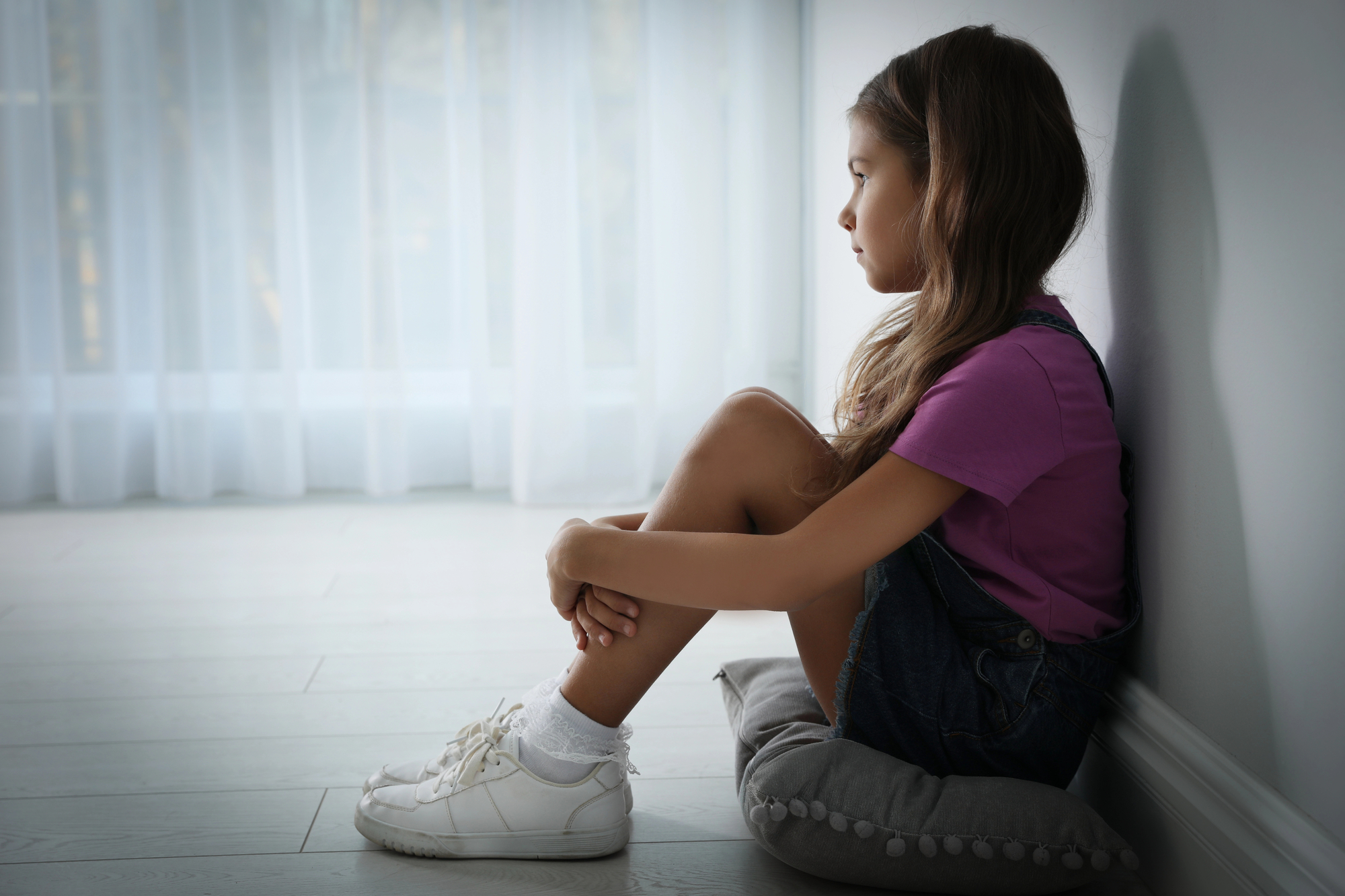 A young girl sits on a cushion near a large window, looking contemplative. She is wearing a purple shirt, denim overalls, and white sneakers. The room is softly lit, with sheer curtains covering the window.