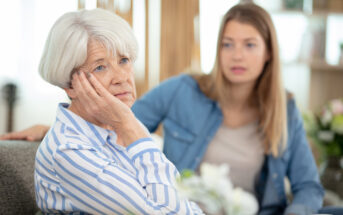 An elderly woman with white hair and a striped shirt sits thoughtfully with her chin resting on her hand. A younger woman with long hair and a denim jacket sits beside her, looking concerned. The setting appears to be a cozy living room.