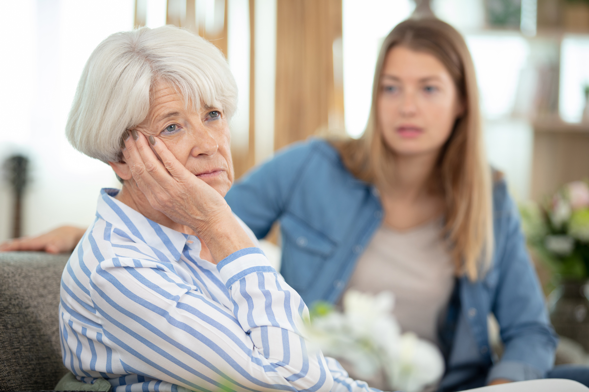 An elderly woman with white hair and a striped shirt sits thoughtfully with her chin resting on her hand. A younger woman with long hair and a denim jacket sits beside her, looking concerned. The setting appears to be a cozy living room.