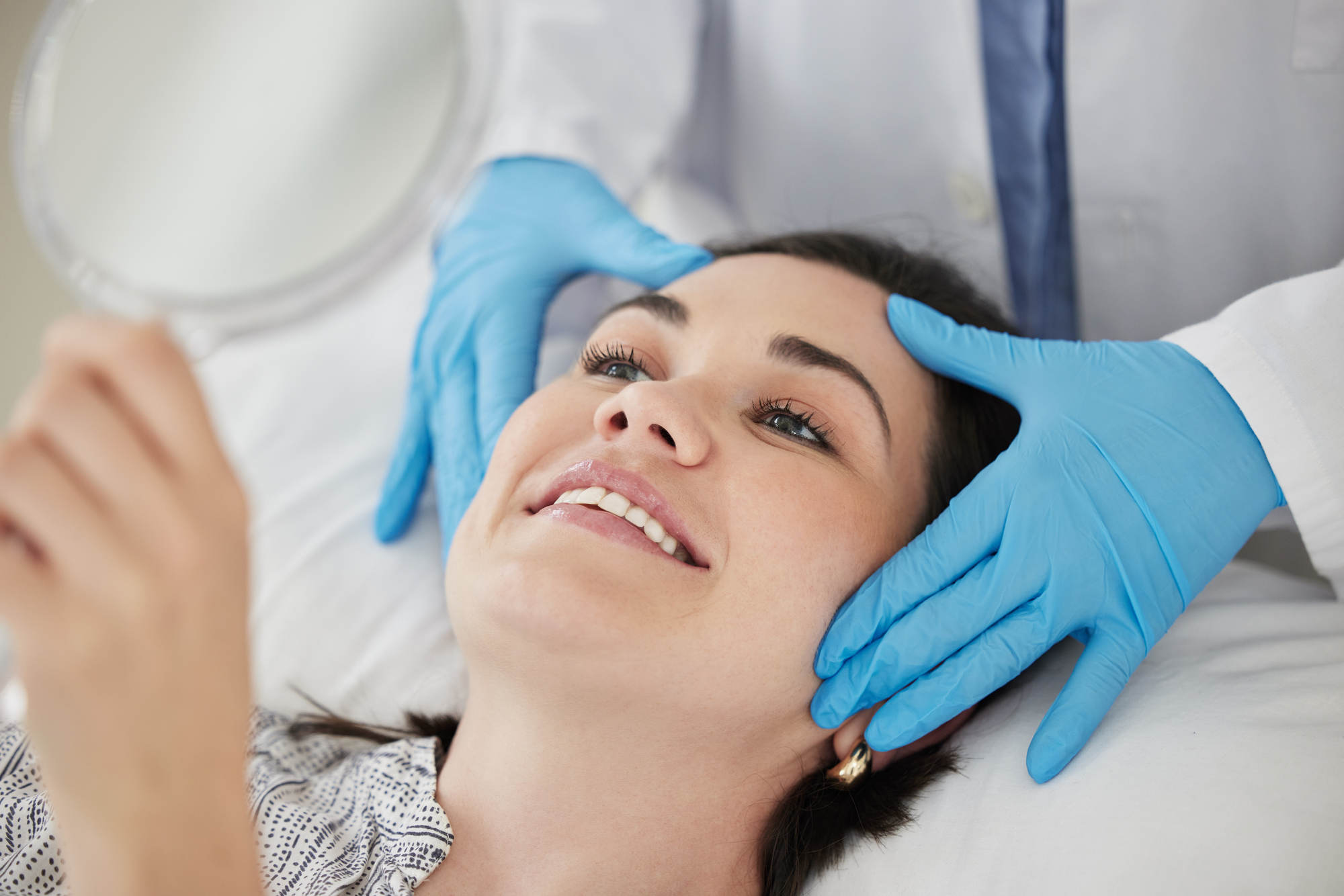 A woman lies on a medical examination table, smiling, while holding a small mirror. A person in a white coat and blue gloves gently examines her face.