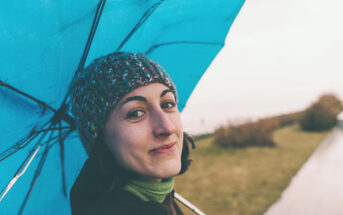 A woman in a knitted hat smiles while holding a blue umbrella. She stands on a path with grass on one side. The atmosphere is overcast but bright.