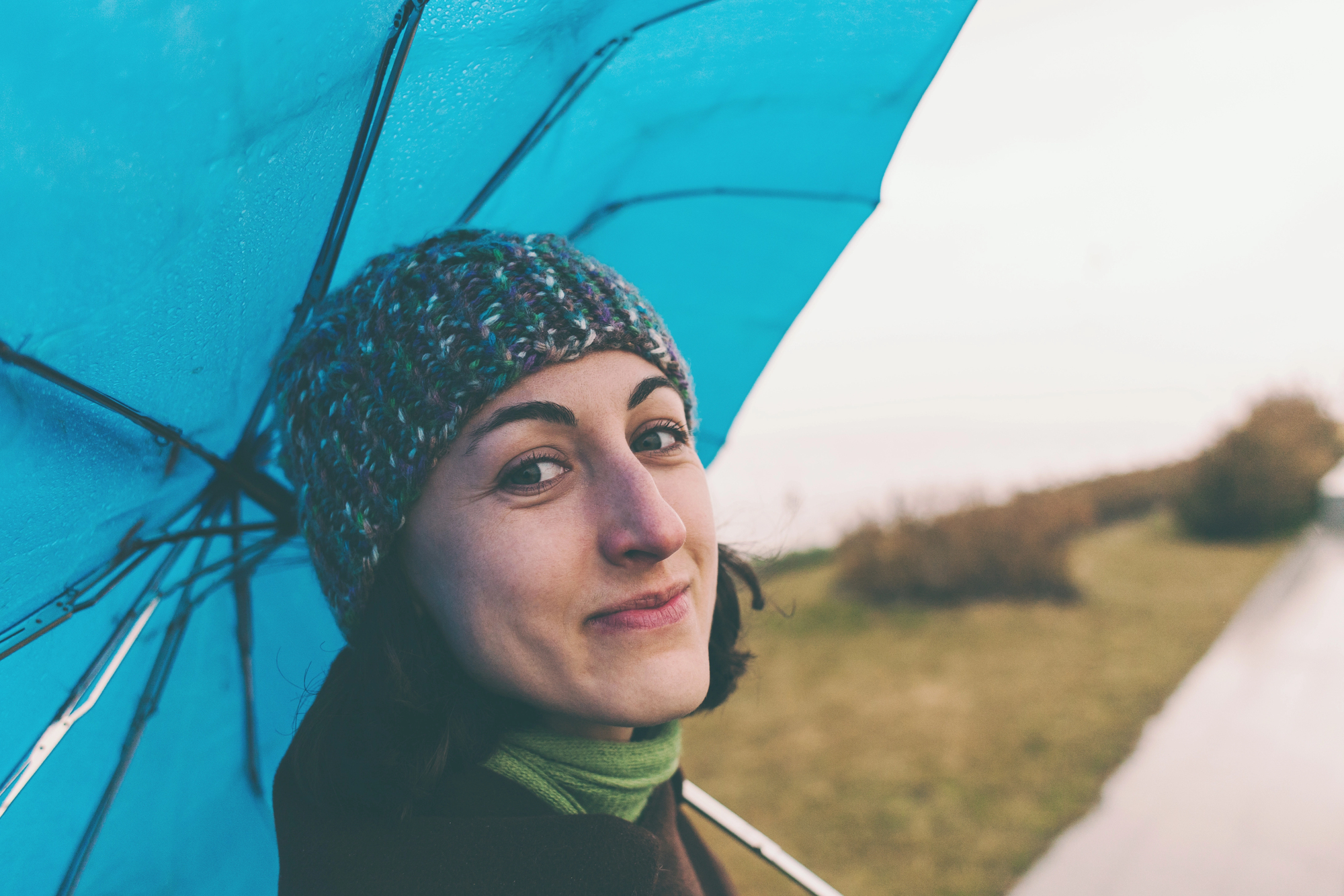 A woman in a knitted hat smiles while holding a blue umbrella. She stands on a path with grass on one side. The atmosphere is overcast but bright.