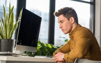 A young man in a brown shirt sits at a desk, staring thoughtfully at a computer screen. Behind him, large windows let in natural light, and greenery, including plants on and around the desk, adds a touch of nature to the scene.