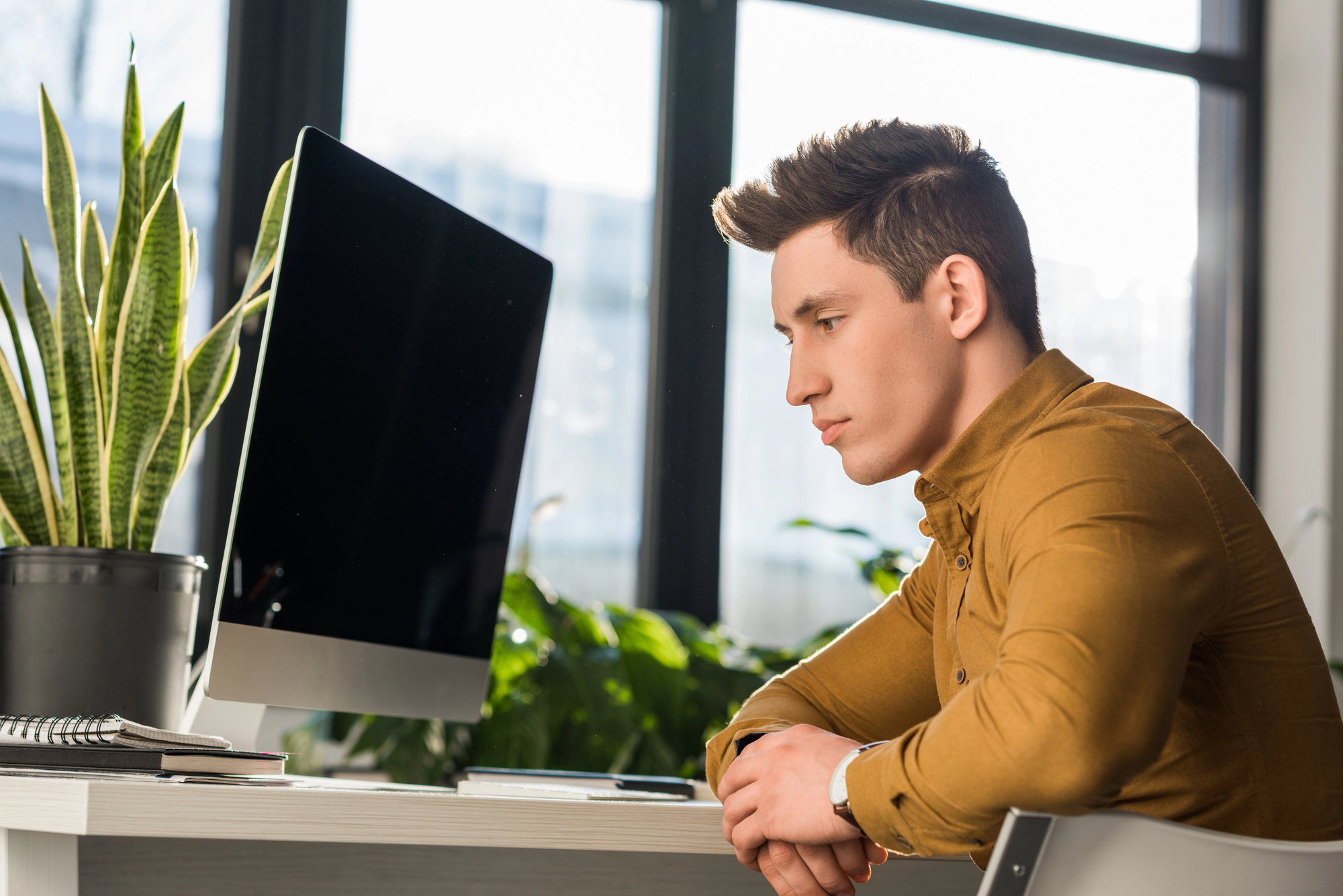 A young man in a brown shirt sits at a desk, staring thoughtfully at a computer screen. Behind him, large windows let in natural light, and greenery, including plants on and around the desk, adds a touch of nature to the scene.