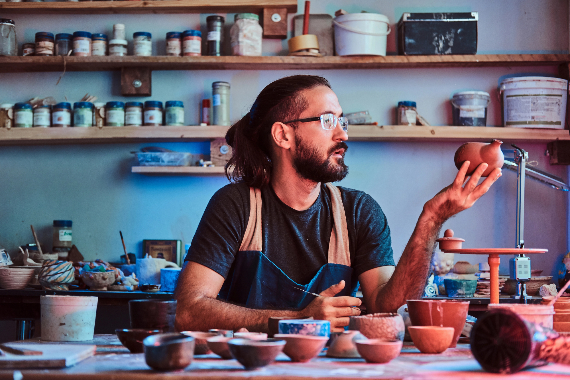 A man with long hair and glasses sits in a pottery studio, examining a small ceramic bowl. The shelves behind him are filled with various pottery jars and tools. A variety of clay pots are placed on the table in front of him.