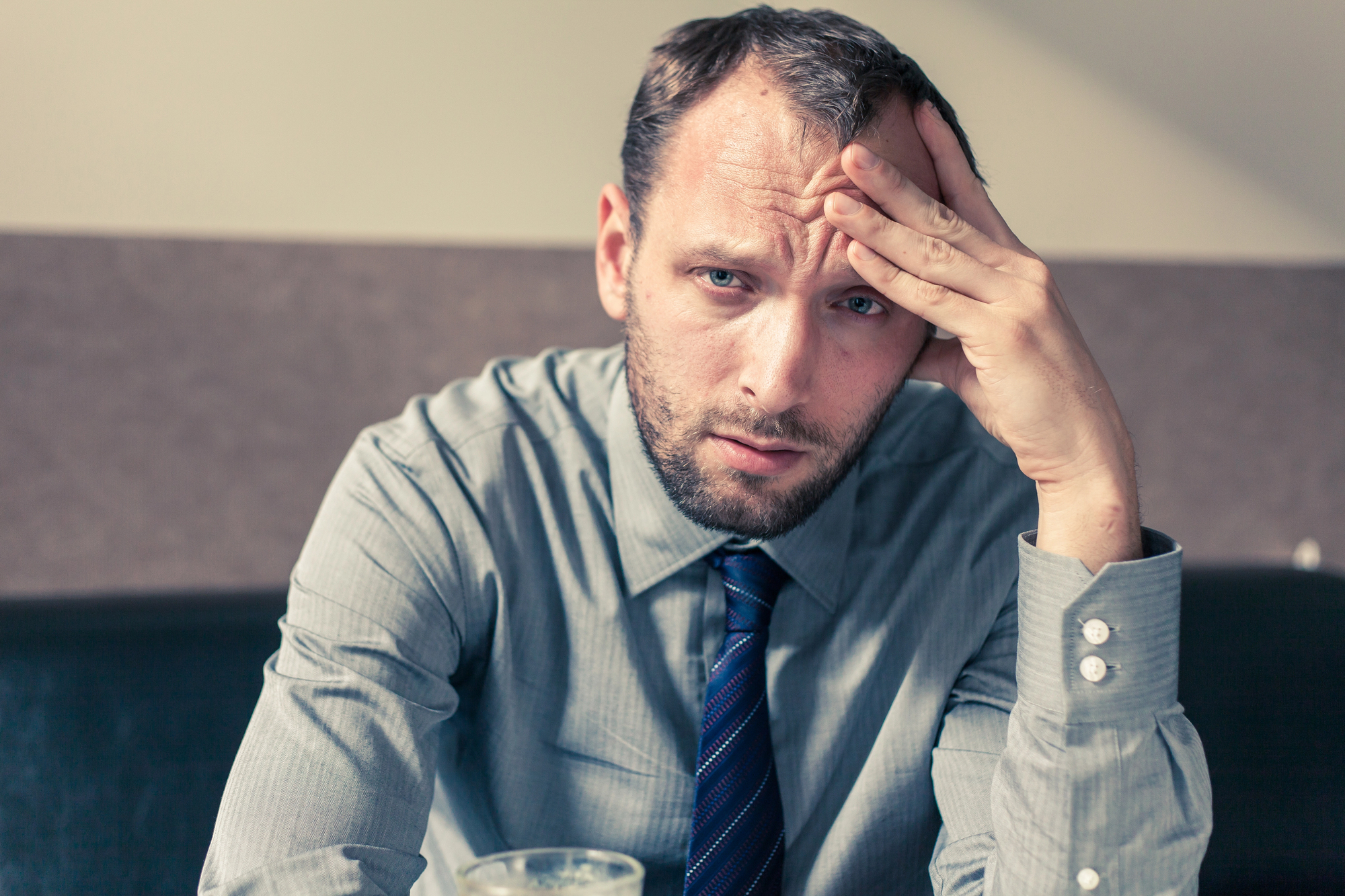 A man in a grey dress shirt and tie sits with his head resting on one hand, looking worried or stressed. He is in an indoor setting, possibly a cafe or office. A drink is partially visible in the foreground.