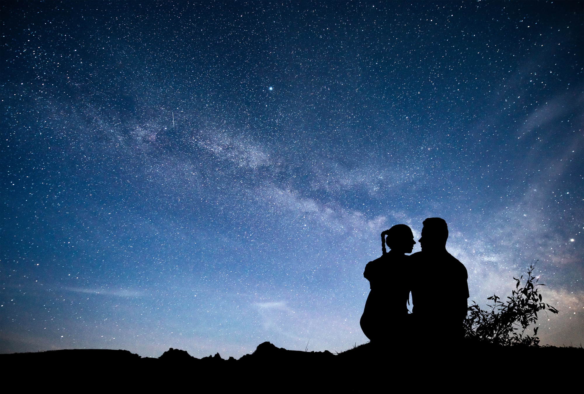 Silhouetted couple sitting and embracing under a vibrant starry sky with a visible Milky Way band, surrounded by a landscape of distant hills and sparse vegetation. The scene conveys a sense of tranquility and wonder.