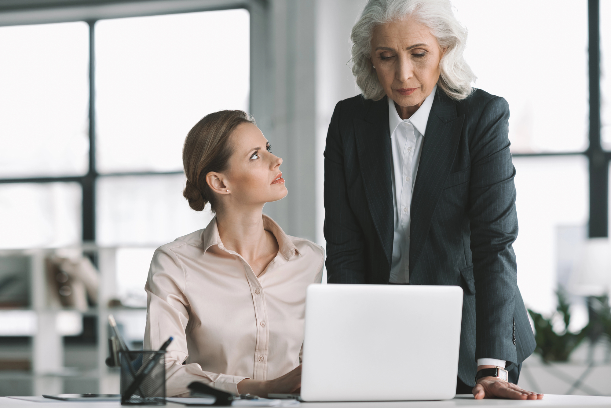 A younger woman seated at a desk looks up at an older woman standing beside her. They are both focused on the laptop in front of them. The setting appears to be an office with large windows in the background.