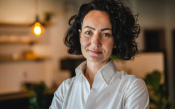 A person with curly hair wearing a white shirt stands indoors with a warm expression. The background is softly lit, featuring a hanging light bulb and blurred shelves.