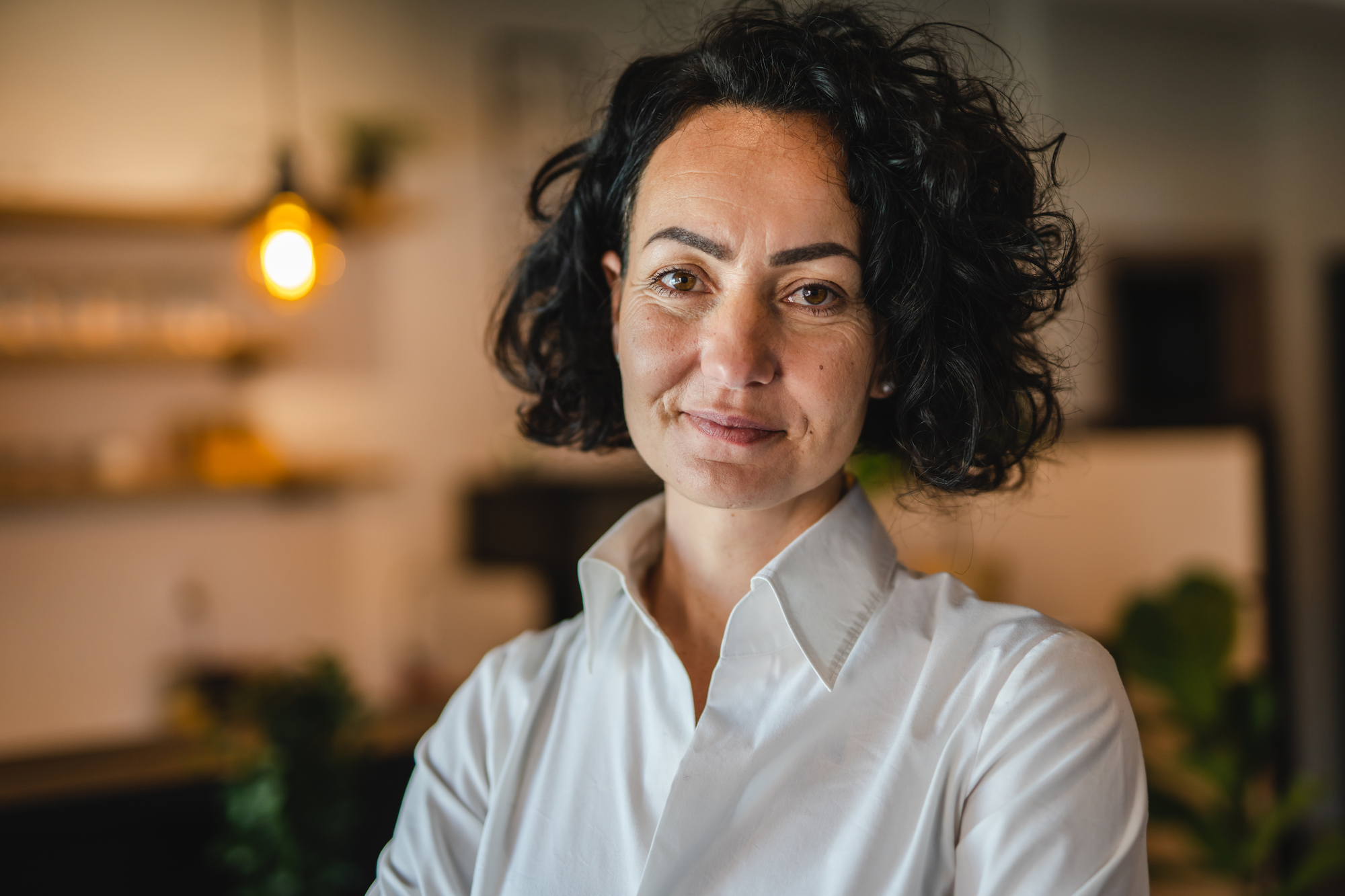 A person with curly hair wearing a white shirt stands indoors with a warm expression. The background is softly lit, featuring a hanging light bulb and blurred shelves.