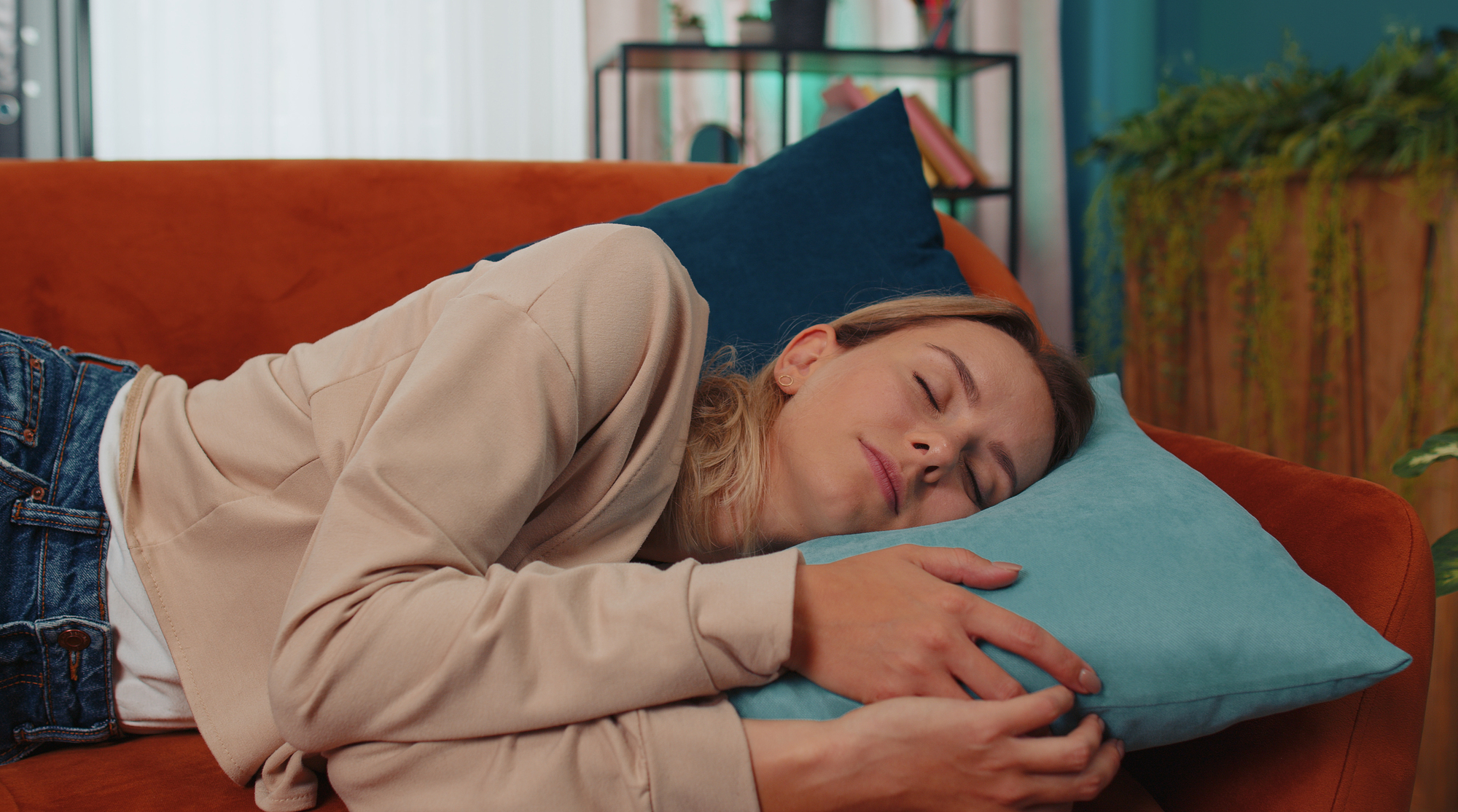 A person with long hair is sleeping peacefully on an orange couch, hugging a light blue pillow. They are wearing a beige top and blue jeans. In the background, there's a shelf with books and plants, and a large plant to the right.