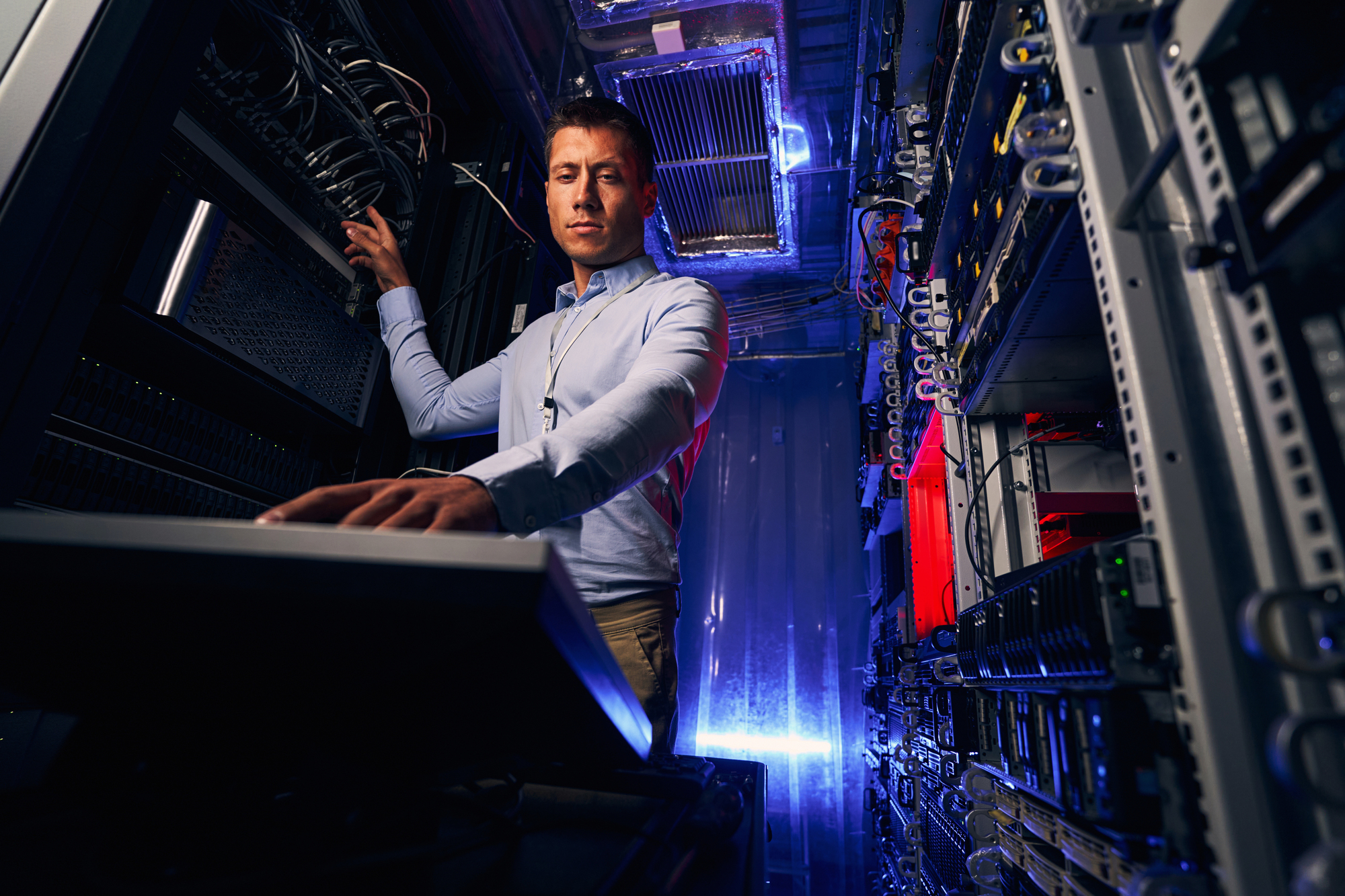 A man in a server room operates a keyboard and monitors cables with focused attention. The room is dimly lit, highlighting the server racks and a blue and red ambient glow.