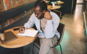 A man sitting in a café, writing in a notebook on a round wooden table. He wears glasses and a checkered shirt. A closed laptop and a coffee cup are on the table. The background shows brick walls and cushioned seating.