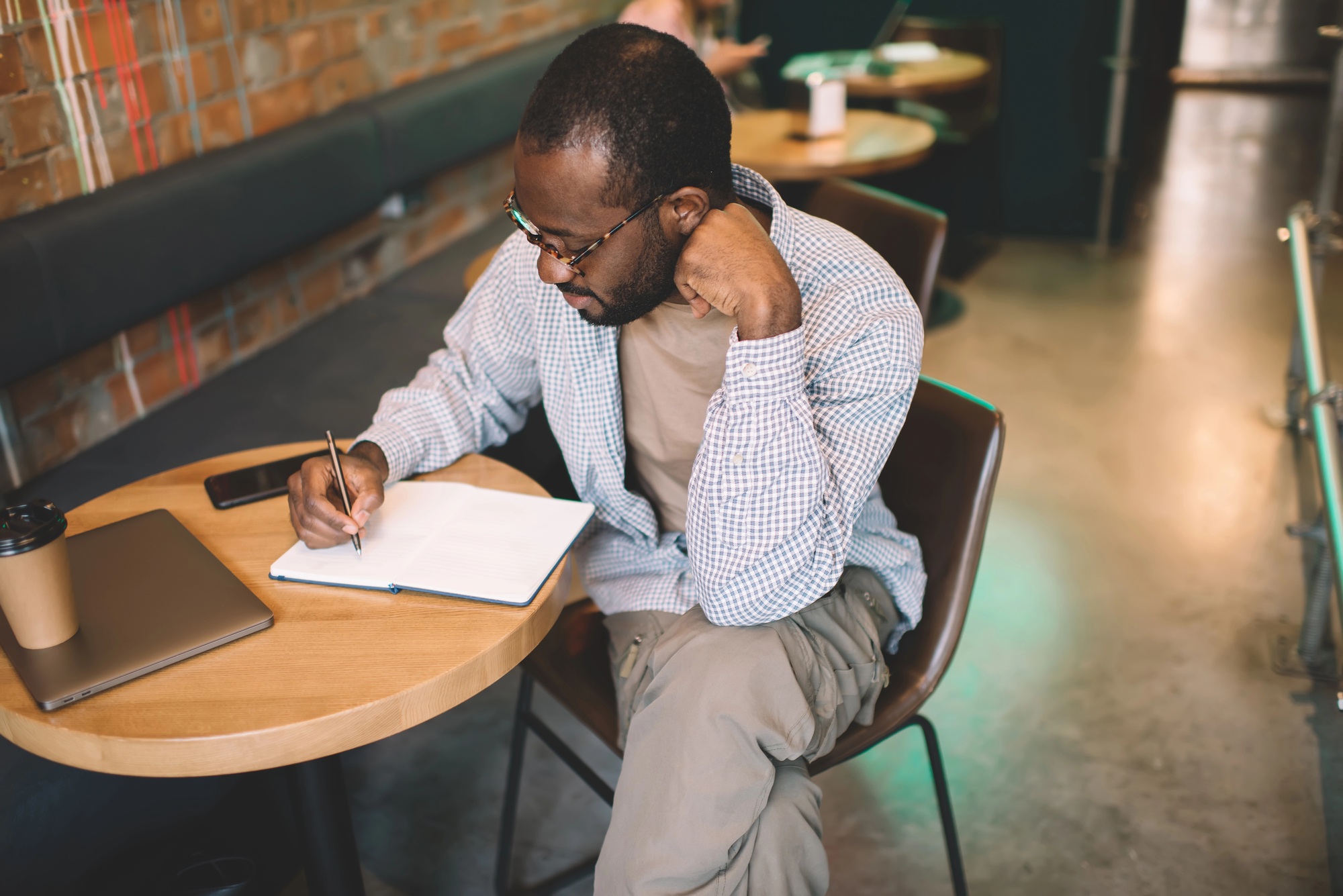 A man sitting in a café, writing in a notebook on a round wooden table. He wears glasses and a checkered shirt. A closed laptop and a coffee cup are on the table. The background shows brick walls and cushioned seating.