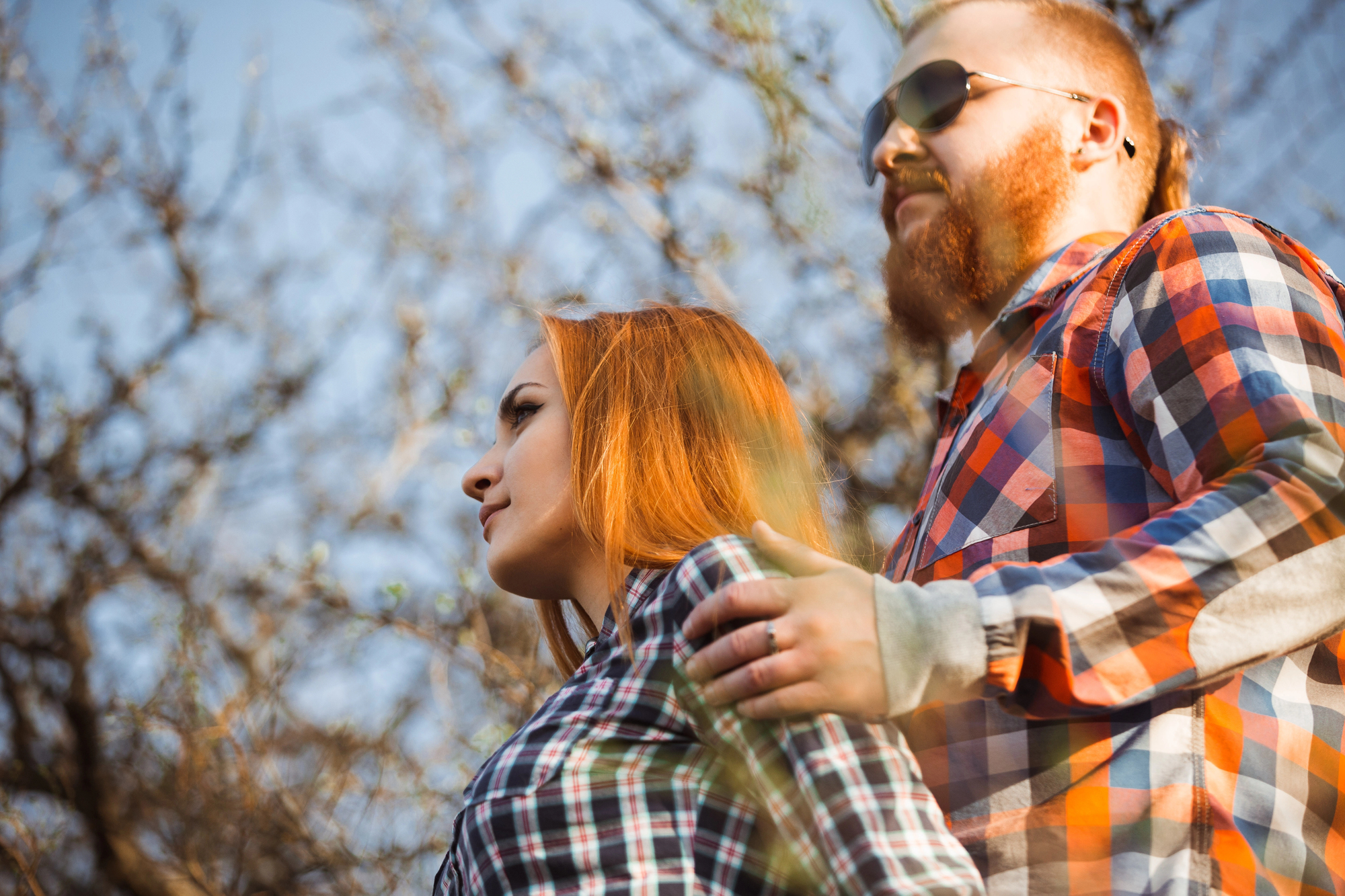 A couple wearing plaid shirts stands close together outdoors. The man with sunglasses and a beard rests his hand on the woman's shoulder. The woman has long red hair, and they are both looking into the distance. Trees and a blue sky are in the background.