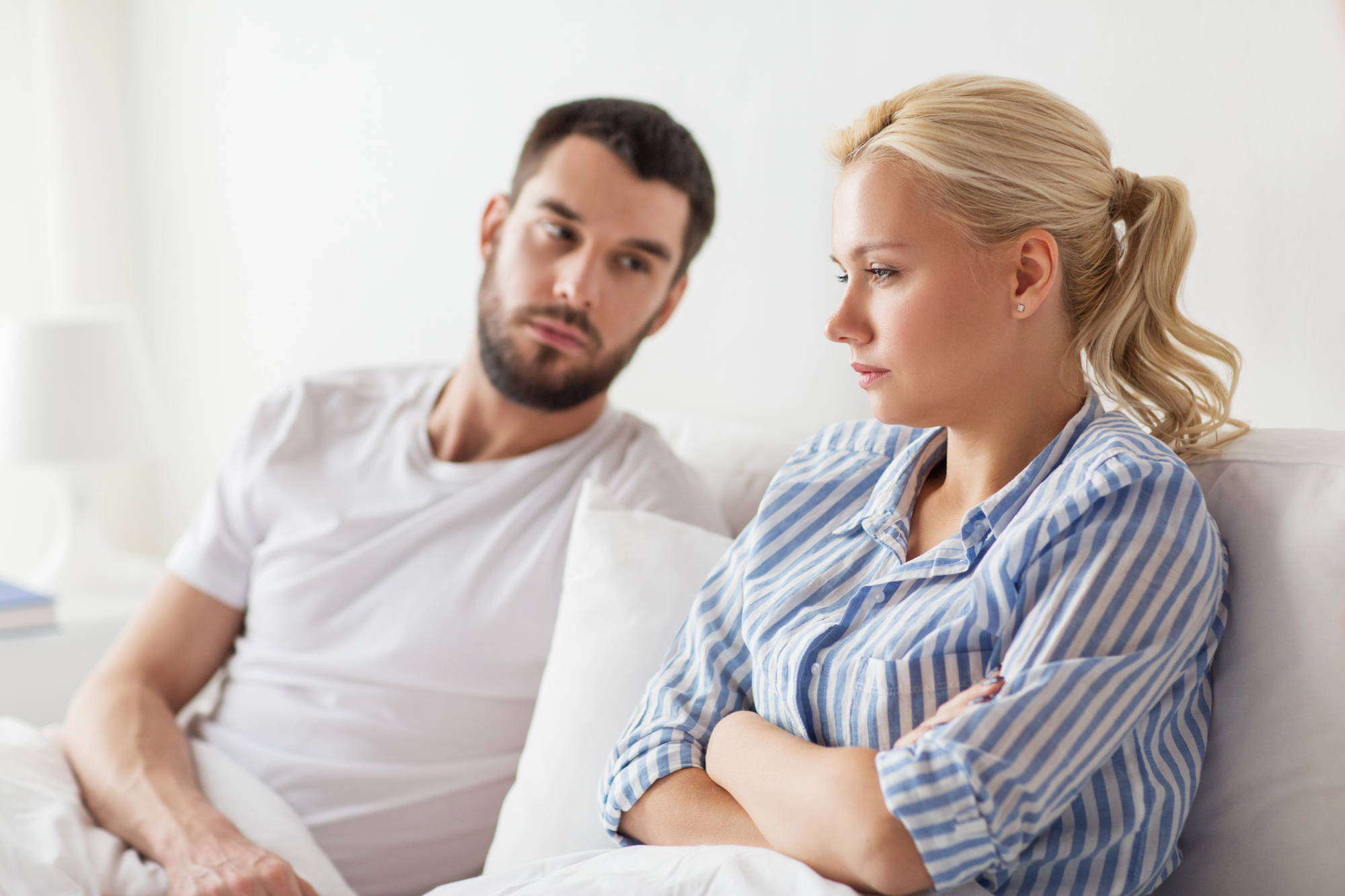 A man and woman sit on a bed, facing each other. The woman, with blonde hair in a ponytail, wears a blue and white striped shirt, looking away with crossed arms. The man, with a beard, wears a white t-shirt and looks at her with a thoughtful expression.