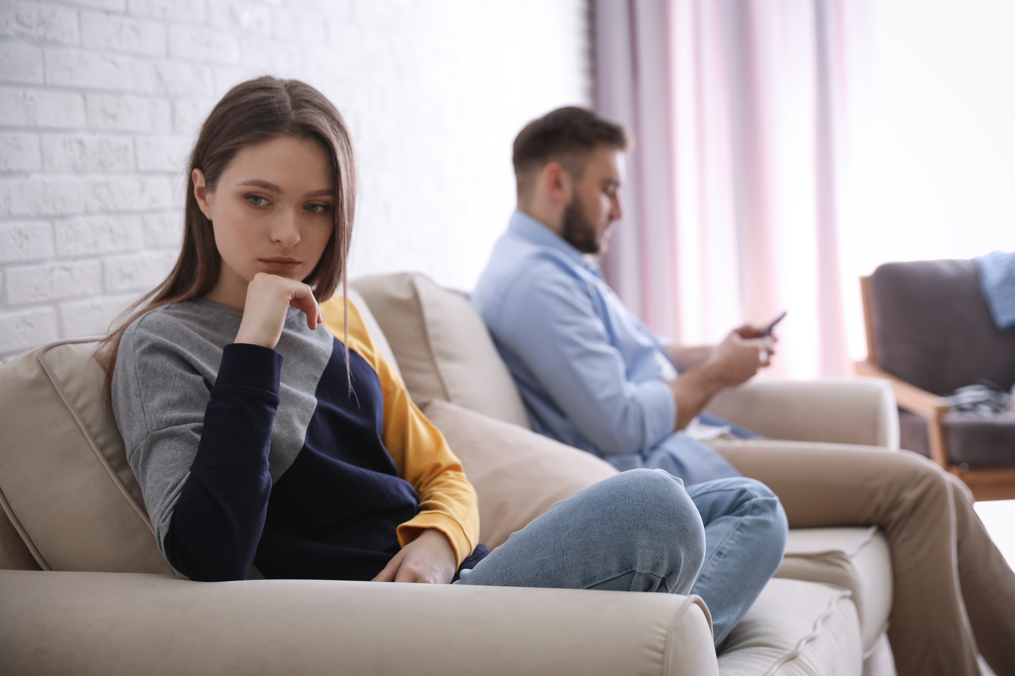 A woman sits pensively on a couch, looking away to her left, while a man sits beside her, engrossed in his phone. The setting appears to be a living room with a light color scheme and natural lighting coming through a window.