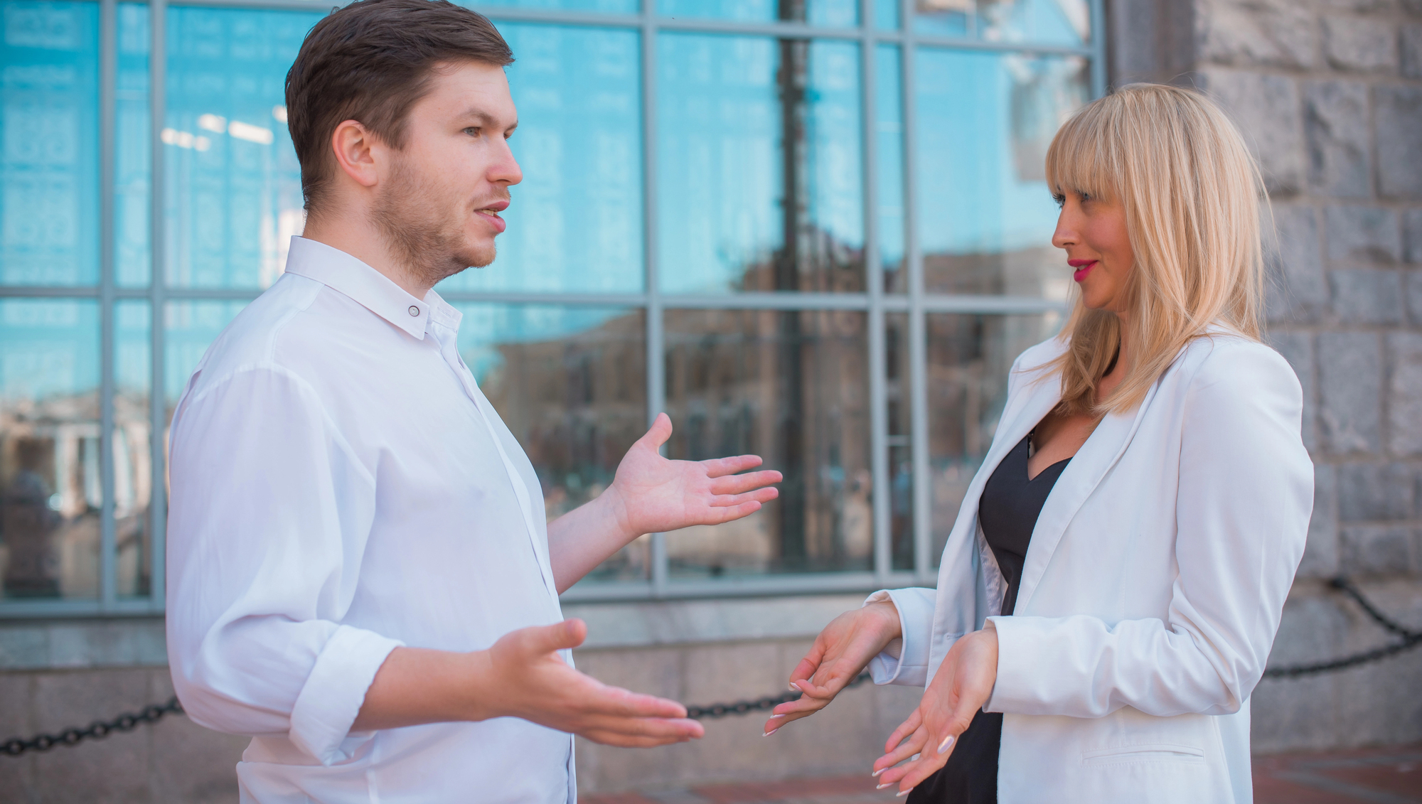 A man and a woman are engaged in conversation outside a building with large windows. Both are wearing white shirts, and the woman has a black dress underneath. Their expressions suggest they are discussing something important.