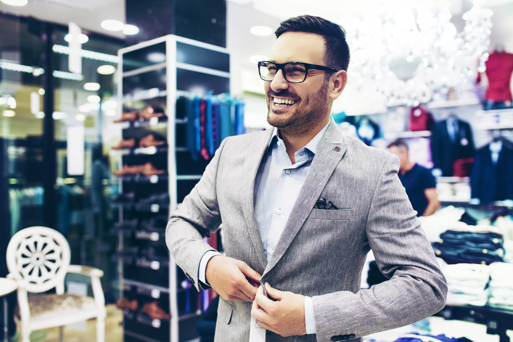 A man in a suit jacket stands smiling in a clothing store. He is adjusting his jacket with one hand. The store is well-lit, with racks of clothes and shoes visible in the background. He is wearing glasses and has short hair.