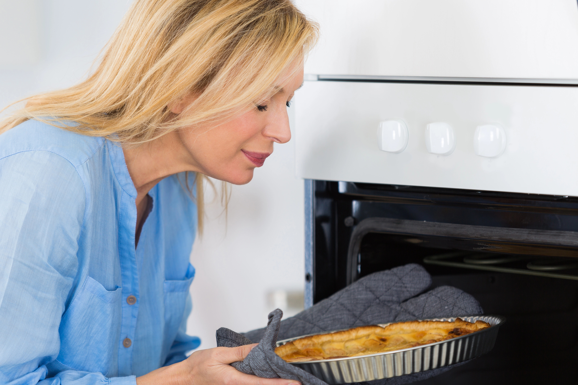 A woman in a blue shirt carefully takes a pie out of an oven using oven mitts. She is smiling as she leans in, enjoying the aroma of the freshly baked pie.